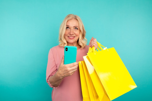 Happy adult woman on the blue wall posing with yellow bags and bright phone.