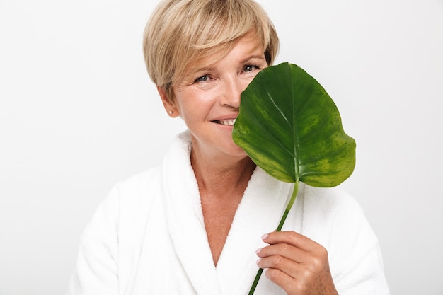  happy adult woman with short blond hair wearing white housecoat holding green leaf isolated over white wall