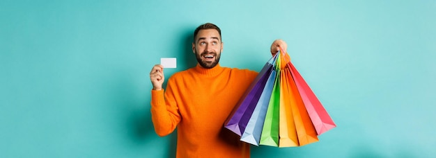 Happy aduly man showing credit card and shopping bags standing against turquoise background