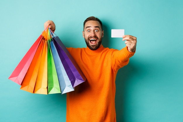 Photo happy aduly man showing credit card and shopping bags, standing against turquoise background