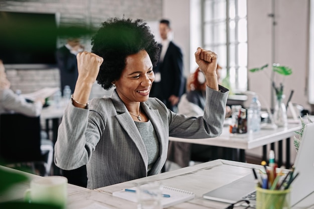 Happy African American businesswoman doing fist pump while working on a computer and celebrating success in the office There are people in the background