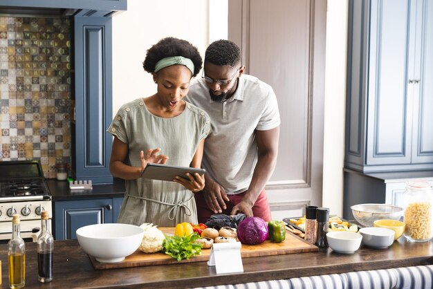 Photo happy african american couple preparing meal together using tablet in kitchen. cooking, healthy lifestyle, recipe, communication, togetherness, food and domestic life, unaltered.