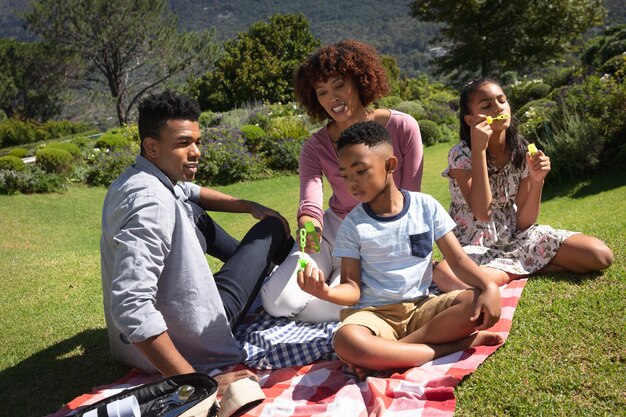 Happy african american couple with son and daughter outdoors, blowing bubbles in sunny garden. family enjoying quality free time together.