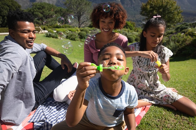 Photo happy african american couple with son and daughter outdoors, blowing bubbles in sunny garden. family enjoying quality free time together.
