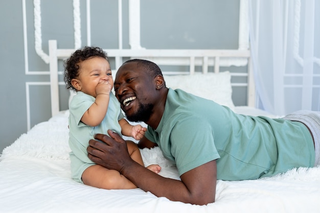 Happy African-American dad hugs his son playing with him on the bed at home, happy family