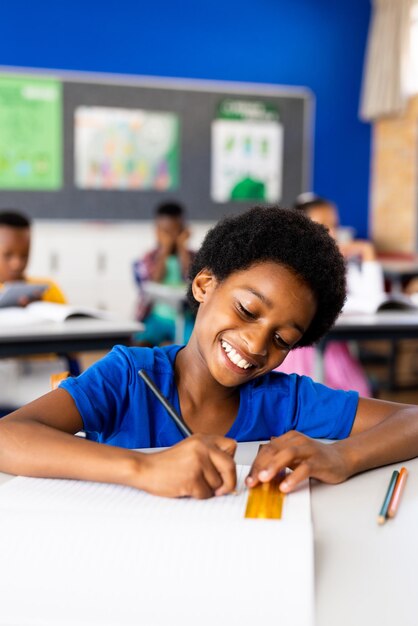 Photo happy african american elementary schoolboy writing in notebook at desk in class copy space