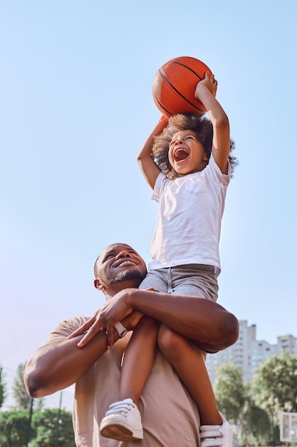 행복한 아프리카계 미국인 아버지가 아이를 들어올리고 농구공을 골대에 넣는 것을 돕습니다.