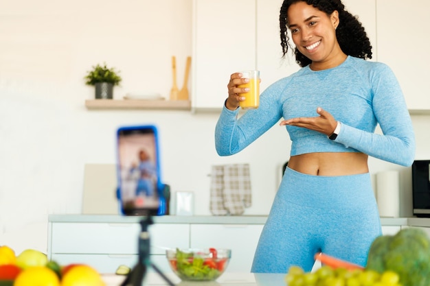 Happy african american fit lady filming video about healthy eating holding glass of juice and talking at phone camera