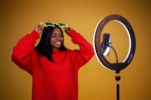 点灯しているリングランプと電話で赤いパーカーを着た幸せなアフリカ系アメリカ人の女の子のブロガー