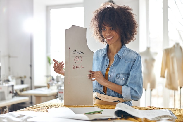 Happy African-American lady dressmaker holds sewing pattern with inscription Size S Black standing at cutting table in spacious workshop