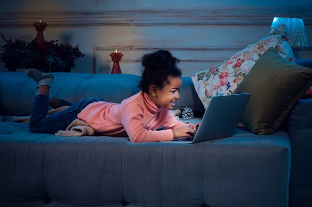 Happy african-american little girl during video call with laptop and home devices, looks delighted and happy. Talking to Santa before New Year's eve, her family, watching cartoons, typing text.