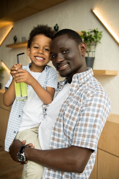 Happy african american man carrying cute little son drinking lemonade and smiling at camera