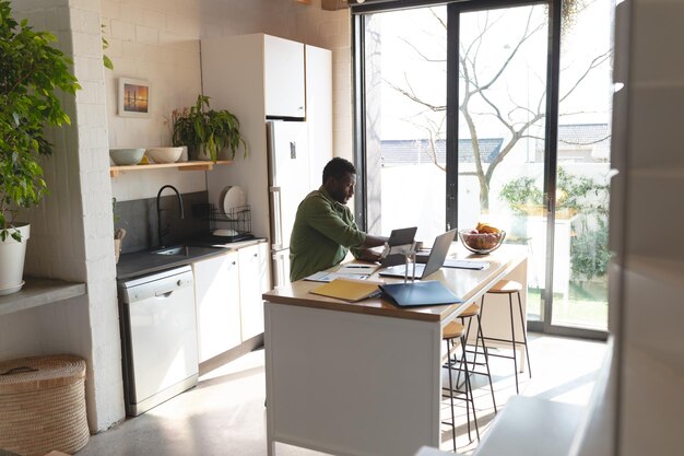 Happy african american man sitting at table in kitchen using laptop. Spending quality time at home alone.