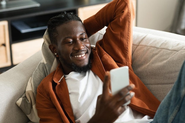 Happy african american man using smartphone browsing internet while lying on couch at home