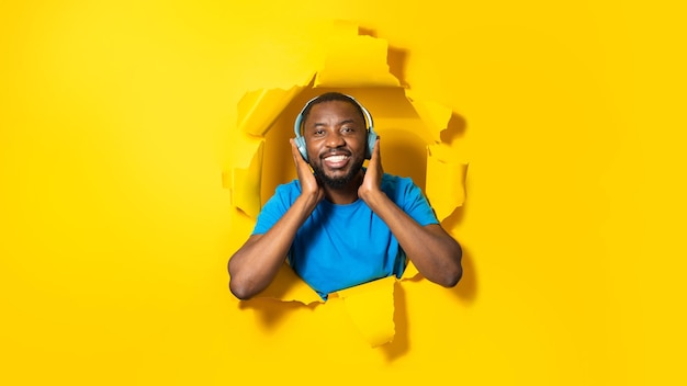 ワイヤレスヘッドフォンを着用し、引き裂かれた黄色い紙の穴からポーズをとる音楽を楽しむ幸せなアフリカ系アメリカ人男性