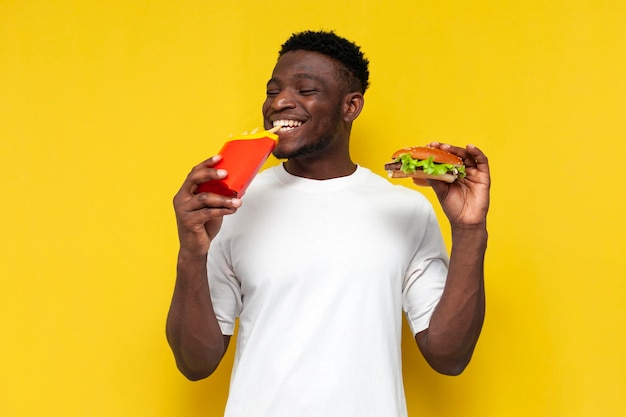 Happy african american man in white tshirt holding big burger and french fries and smiling