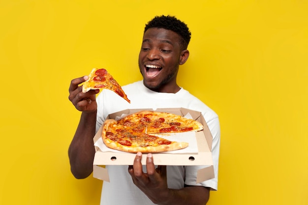 Happy african american man in white tshirt holds box of pizza and rejoices on yellow background