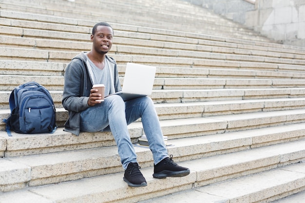 Happy african-american man with laptop and take away coffee sitting on stairs. Casual student preparing for exams with computer in university campus. Education and technology concept.