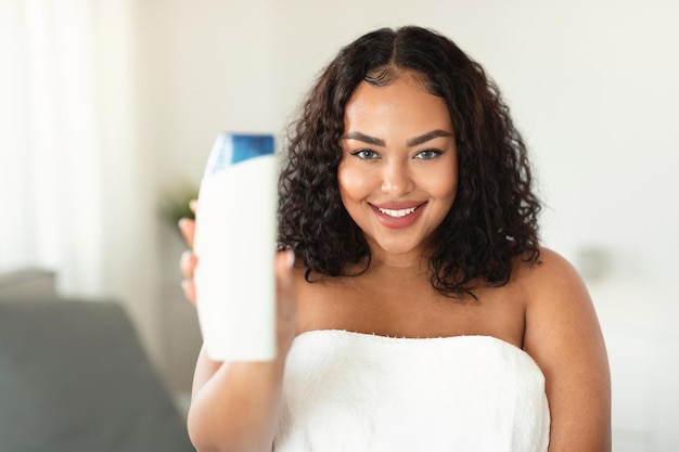 Felice donna afroamericana oversize che mostra una bottiglia di gel doccia che pubblicizza un prodotto cosmetico per il corpo e la cura dei capelli