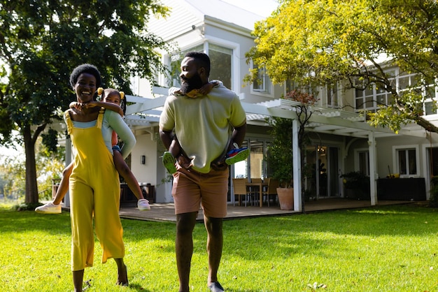 happy african american parents piggybacking playful son and daughter while walking in yard. unaltered, lifestyle, family, love, togetherness, childhood, house, playing and enjoyment.