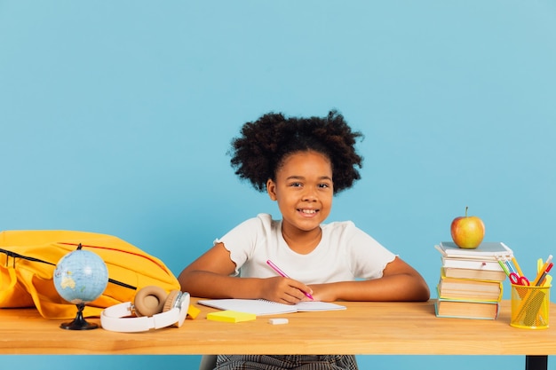 Happy African American schoolgirl doing homework at desk in class on blue background