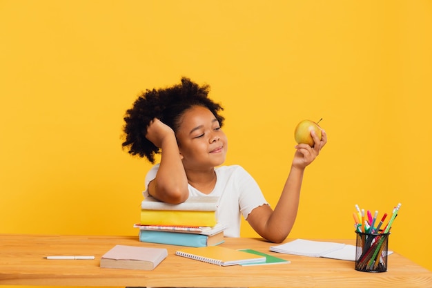 Happy African American schoolgirl doing homework and holding apple for lunch while sitting at desk