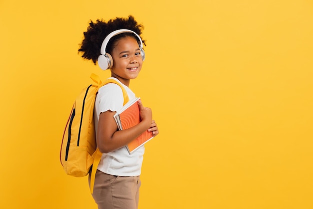 Счастливая африканская школьница в наушниках с рюкзаком, держащая тетради на желтом фоне
