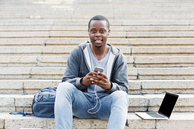 Happy african-american student sitting on stairs and listening to music on his smartphone outdoors, having a rest in the university campus with copy space. Education concept, copy space