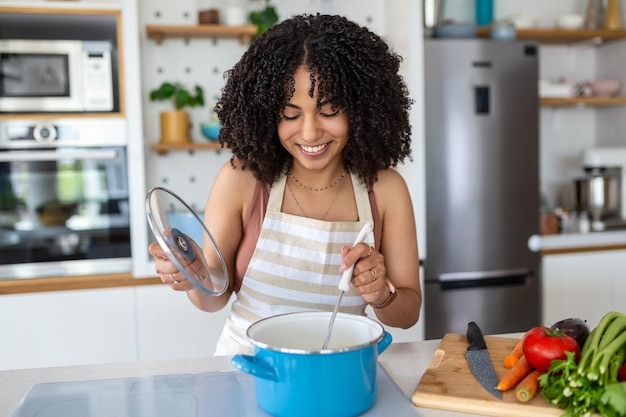 Felice donna afroamericana cucinare degustazione cena in una pentola in piedi nella cucina moderna a casa casalinga preparare cibo sano sorridente alla fotocamera famiglia e nutrizione dieta ricette concetto