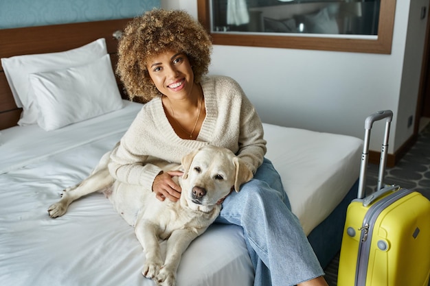 행복한 아프리카계 미국인 여성이 애완동물 친화적인 호텔 방에서 수하물 근처에 라브라도르를 안고 있습니다.