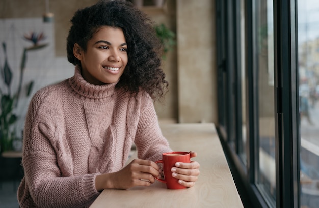 Happy African American woman drinking coffee, looking through the window, stay home