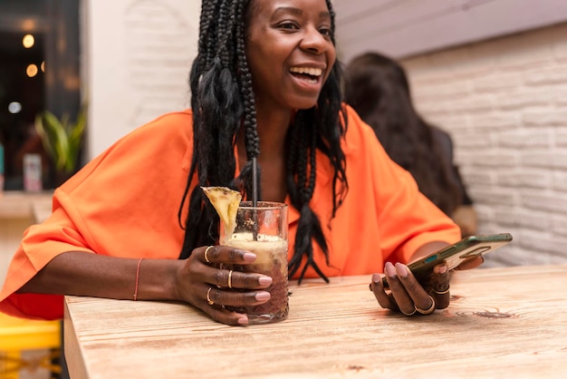 Happy African American woman holding a drink at the bar while uses mobile phone