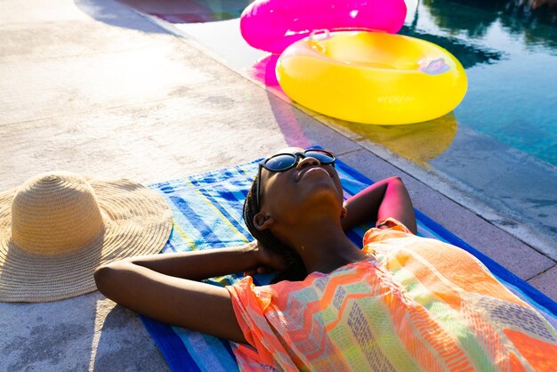 Happy african american woman lying on towel and sunbathing next to swimming pool in garden. Lifestyle, free time and vacation, summer, sunshine, unaltered.