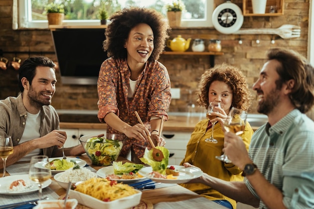 Felice donna afroamericana che serve insalata ai suoi amici mentre pranza insieme a casa