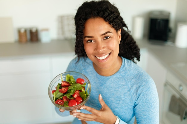 Photo happy african american woman in sportswear eating healthy salad after domestic workout sitting in kitchen and smiling