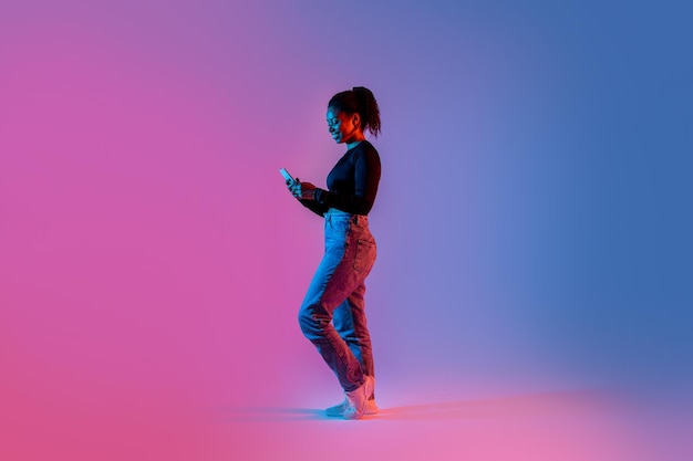 Happy african american woman texting on cellphone standing in colorful neon light full length free space