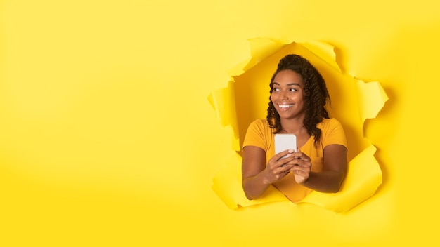 Happy african american woman with cellphone smiling and looking aside at free space through hole in torn paper panorama