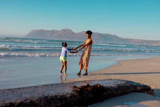 행복한 아프리카계 미국인 젊은 여성이 딸의 손을 잡고 맑은 하늘을 배경으로 해변에서 즐거운 시간을 보내고 있습니다.