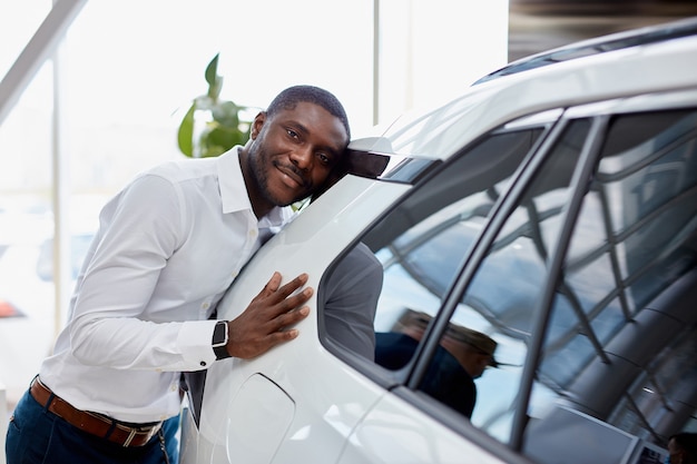 L'uomo d'affari africano felice abbraccia la sua nuova auto di lusso bianca in concessionaria