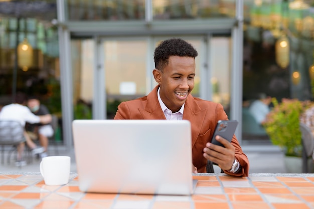 Happy African businessman outdoors in coffee shop using laptop computer and mobile phone