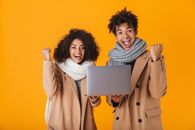 Happy african couple wearing winter clothes standing isolated, holding laptop computer, celebrating success