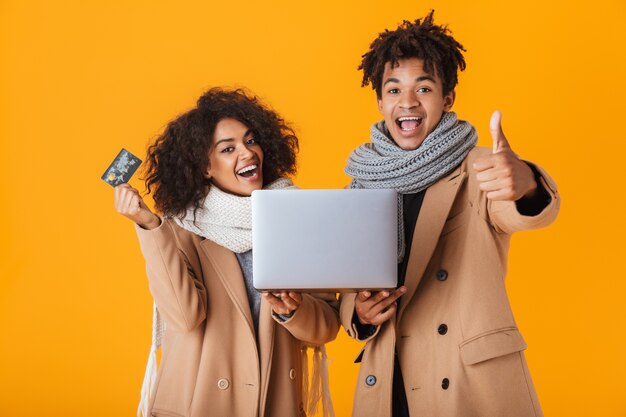 Happy african couple wearing winter clothes standing isolated, holding laptop computer, showing plastic credit card, celebrating