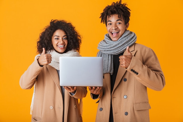 Happy african couple wearing winter clothes standing isolated, holding laptop computer, thumbs up
