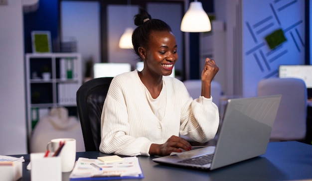 회사 사무실에서 초과 근무를 하는 노트북에 대한 좋은 소식을 듣고 있는 행복한 아프리카 프리랜서