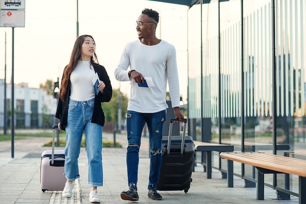 Happy african guy with his smiling asian woman-mate holding passports with tickets and carrying suitcases near airport.