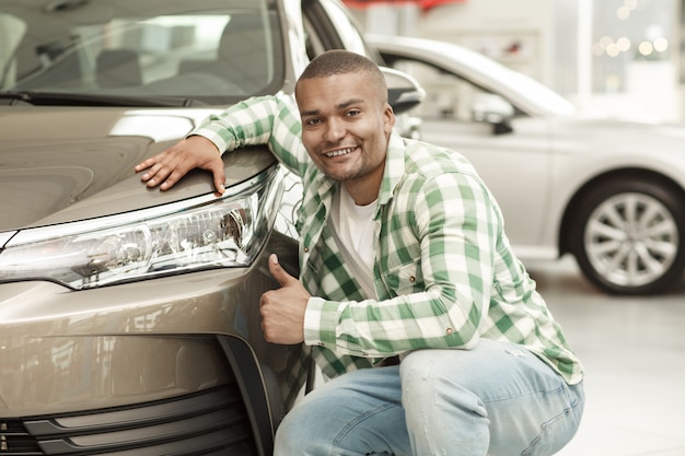 Foto uomo bello africano felice che mostra i pollici su che posano vicino alla sua nuova automobile al concessionario