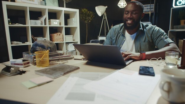 Happy african man flirting online in dark coworking Successful african american businessman typing messages on computer in hipster office Cheerful afro guy communicating on laptop indoors