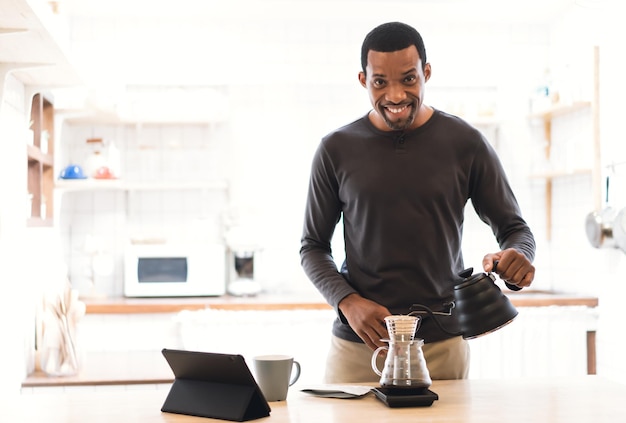 Happy African middle age man learning online making coffee class via digital tablet and looking at camera poured hot water from the kettle into the glass jar to make coffee in modern kitchen at home