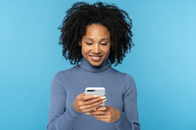 Happy African millennial woman with curly hair chatting in social media, using mobile phone isolated