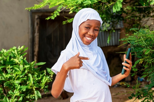 happy African Muslim schoolgirl pointing at a cell phone while wearing a white hijab